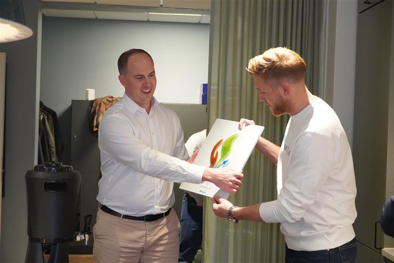 To personer står indendørs og tager imod en pris sammen. Den ene overrækker en plakette med Indutrade Sustainability Awards-symbolet til den anden.